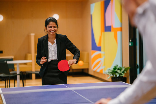 Portrait of a young and attractive Indian Asian woman in a suit playing table tennis with her colleague in the office during a break. She is having lots of fun and is smiling broadly.