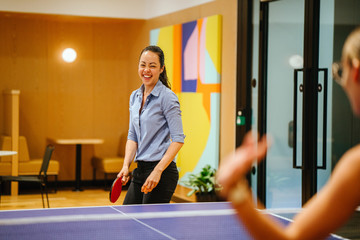 Portrait of a young and attractive Chinese Asian woman professionally dressed in business attire playing table tennis in the office with a colleague. She is smiling as she enjoys the break. - obrazy, fototapety, plakaty