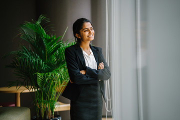 Portrait of a young and attractive Asian Indian woman in a suit during the day standing by a window. She smiles as she looks at the view.