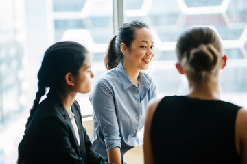 Image of a young Asian Chinese manager chatting in their office with her team. As she listens to her colleague speaking about their new project, she smiles back.