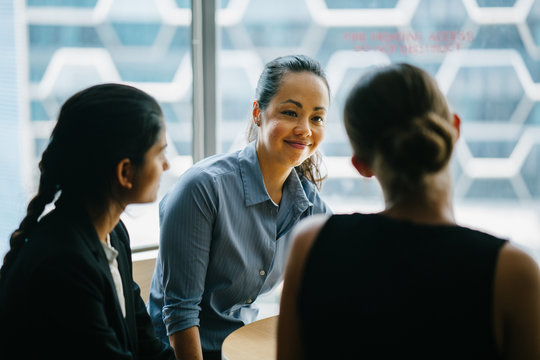Image of a young Chinese Asian woman manager chatting with her team in their office. She smiles back as she listens to her colleague talking about their new project.
