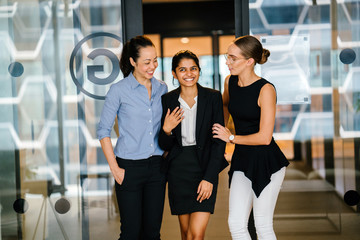 Portrait of a group of professional women standing and smiling in their office lobby against a glass door. They're all dressed professionally and competently.