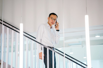  A Chinese Asian executive is walking down the stairs while using his smartphone. He is talking to...