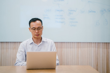 Portrait of relaxed Chinese Asian businessman sitting at a desk while typing on his laptop. He is wearing a professional outfit and is looking focused doing his task.