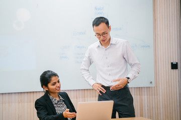 A portrait of two professional business people talking inside a conference room. They are deep in conversation while using the laptop as a reference. Both of them are dressed professionally.