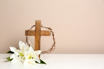 Wooden cross, crown of thorns and blossom lilies on table against color background, space for text