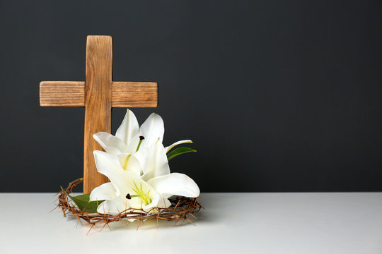 Wooden cross, crown of thorns and blossom lilies on table against color background, space for text