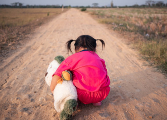Behind of Asian little girl is holding doggy doll and standing at the entrance to the rice fields near the house in sunset.