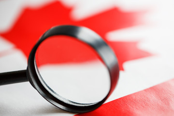 Obraz na płótnie Canvas The flag of CANADA is looked through a magnifying glass. Espionage and surveillance of Canada. Monitoring the status of Kanatsev The concept of the danger of theft of military terrorism