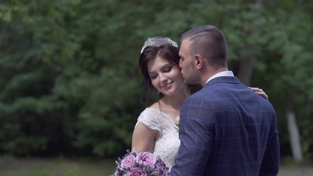 handsome groom in blue checkered jacket kisses pretty bride in tiara scratching open shoulder slow motion
