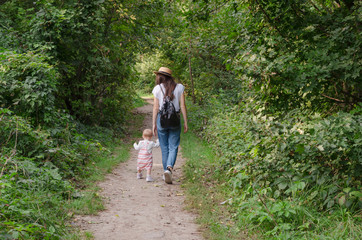 Obraz premium Beautiful brunette female in straw hat jeans, white t-shirt and a small backpack with his little cute child daughter playing together in a park