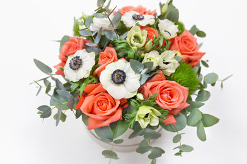 Delicate floral arrangement of roses, anemones (white, pink, orange) and green eucalyptus leaves in a round white cardboard box on a bright background