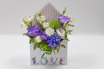 Delicate floral arrangement (gift) of fresh flowers in a box in the form of an envelope with the inscription Love, on a light background. (eustoma, leaves. Colors: purple, lilac, white, green).