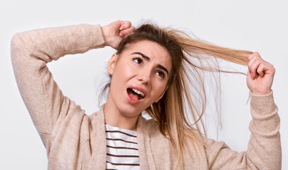 Close up image of upset young woman trying comb unruly hair pulling strands with raised hands screaming from pain and discomfort,  posing over white background. People, health and care concept