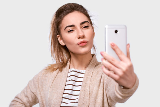 Studio close up studio shot of a pretty young European woman in casual clothes sending air kiss to his boyfriend while standing and taking a selfie isolated over white studio background.