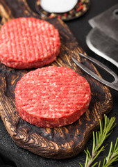 Fresh raw minced homemade farmers grill beef burgers on vintage chopping board with spices and herbs and meat hatchet, fork and knife on black board.