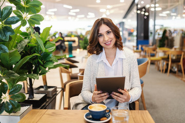 Gorgeous smiling brunette dressed casual using tablet and sitting in cafeteria. On desk coffee and glass of water. Woman looking at camera.