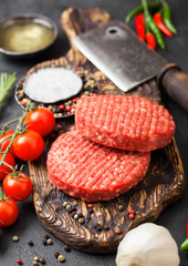 Fresh raw minced homemade farmers grill beef burgers on vintage chopping board with spices and herbs and meat hatchet on black board. Oil, garlic and tomatoes.