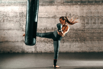 Dedicated strong brunette with ponytail, in sportswear, bare foot and with boxing gloves kicking...