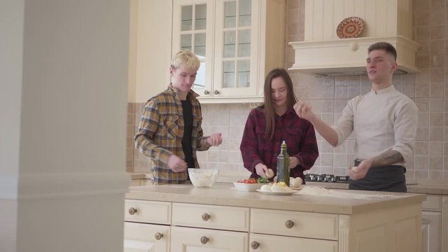 Young pizza maker juggles balls of dough in the kitchen, demonstrating his skills to his friends. Smiling man and woman unsuccessfully trying to repeat his actions. Pizza cooking art