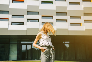 Beautiful hipster girl is standing near the modern architecture building. Stylish business woman walking near the fashionable lines, glass and windows. Green grass lawn. Office worker break.