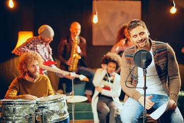 Man singing and sitting on chair while his band playing instruments in background. Home studio...