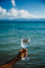 Aquamarine sea background with blue sky and white clouds skyline wine glass with brilliant liquid natural light tanned female hand and shadows and caustic landscape