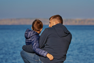 Boy is sitting on his father’s knees on the bank of the lake. They are happy to spend time together.