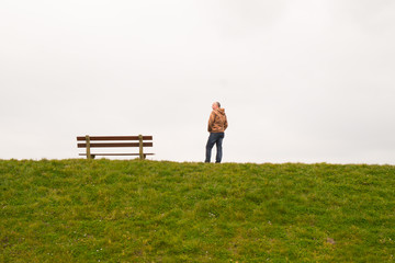 Naklejka na ściany i meble A single person standing next to a single empty wooden bench on the horizon