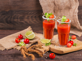 Two glasses of fresh organic tomato juice decorated with raw tomatoes, cucumber and leaves on a rustic wooden cutting board