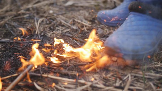 The woman in galoshes extinguishes the flaring dry grass stepping on a fire.