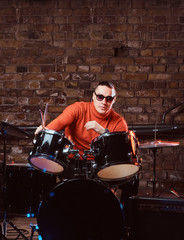 Fototapeta na wymiar Young stylish musician in sunglasses sits behind the drum set against a brick wall, thoughtfully looking at the camera. Perform in a night club