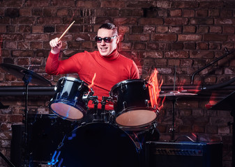 Young stylish musician in sunglasses emotionally playing drums against brick wall background
