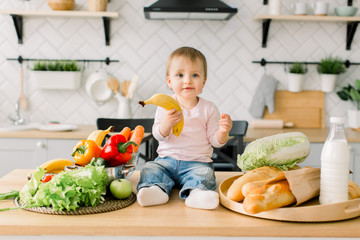 Beautiful little girl one year old, sitting on a table in the kitchen among fruits and vegetables and holding a banana in her hand. Modern kitchen on the background