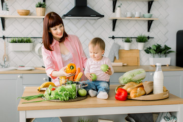 mother and kid daughter cooking in the kitchen . Mom holds pepper in her hand and her daughter holds the apples and eats them