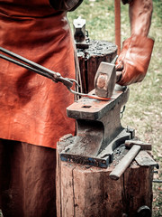 The blacksmith in the production process of metal products handmade on the openair workshop. Blacksmith strikes with a hammer on metal. Hands of a blacksmith in leather gloves with red-hot billet