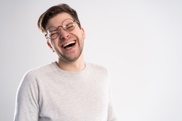 Portrait of young Caucasian guy enjoys funny story or joke, laughs joyfully, has toothy smile, wears white t shirt in one tone with background. Happiness and youth concept.