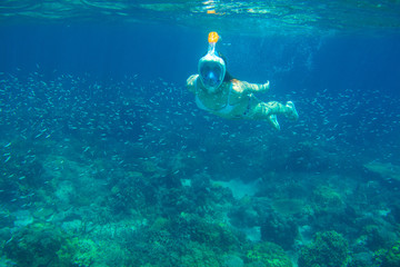 Girl in snorkeling mask underwater photo in blue sea. Diving in fish school. Woman in full-face snorkeling mask.