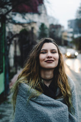 Naklejka premium Smiling young caucasian model standing on the sidewalk. Blue wet asphalt after autumn rain. Early evening. Urban concept. Headlight of the car appearing behind the corner of the street. Cinematic scen
