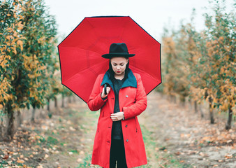 Woman in red coat and with umbrella between trees in apple garden at autumn season. Beautiful...