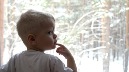 In a slow motion video shoot, a small child (boy) looks out the window on the winter day as snowflakes fall. Concept of: Winter day, Cold day, Mood winter, Snow clapping.