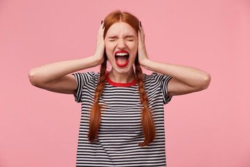 Angry young red-haired girl in stripped t-shirt, keeps closed eyes, closes her ears with palms, showing gesture of ignore, demonstrates loud scream or shout, over pink wall