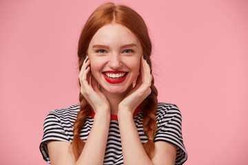 Close up of gorgeous pretty red-haired girl with two braids keeps hands near her face and smiling lively with red lips, showing white healthy teeth, isolated on pink background