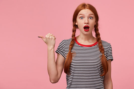 Shocked red-haired girl with two braids pointing with thumb to the left side on blank copy space for your text, openes mouth widely saying OMG, draws attention, isolated on pink wall