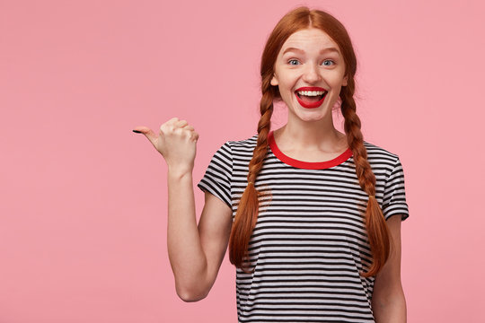 Overflowing with positive emotions red-haired girl with two braids pointing with thumb to the left side on blank copy space, openes mouth widely in excitement, with red lips, isolated on pink wall