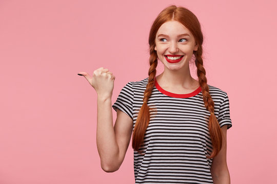 Happy excited red-haired girl with two braids pointing with thumb to the left side on blank copy space and looks there with interest, smiling widely, draws attention, with red lips, on pink wall