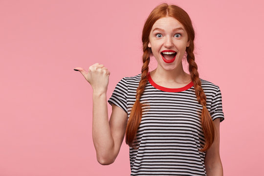 Excited red-haired girl with two braids pointing with thumb to the left side on blank copy space, openes mouth widely in surprise, draws your attention, with red lips, isolated on pink wall