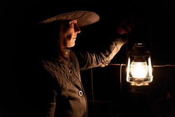 Cowgirl wearing hat walking in dark by lantern light beside farm fence in field © BLM Imaging