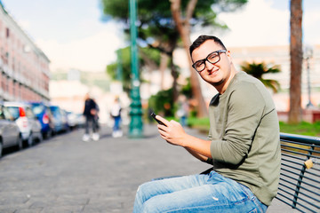 Young man with smartphone sitting on city street bench, copy space. Portrait of young man using mobile phone and looking at screen outdoor, side view. People, technology and travel concept