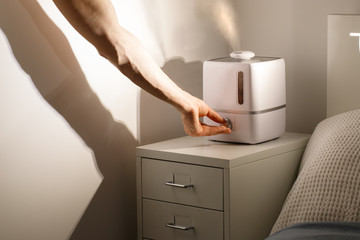 Hand turn on aroma oil diffuser on the bedside table at night at home, steam from the air humidifier. Ultrasonic technology, increase in air humidity indoors, comfortable sleeping conditions. - obrazy, fototapety, plakaty